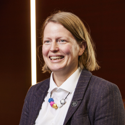 A white woman with blonde hair wearing a white shirt, grey jacket and colourful necklace